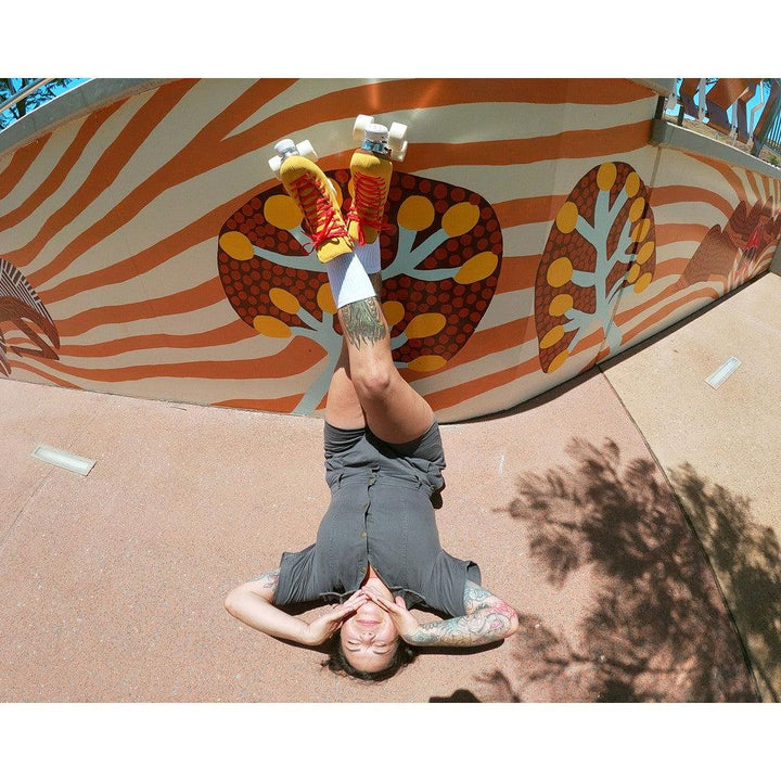 roller skater laying on her back with legs crossed wearing Chuffed Skates yellow roller skates with aboriginal painting wall behind